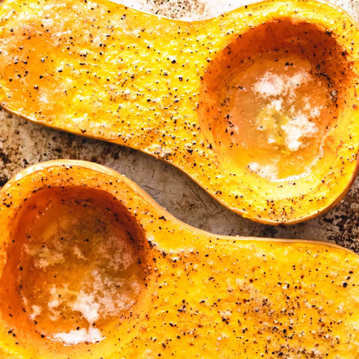 Two baked butternut squash halves on a baking sheet with melted butter and pepper.