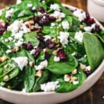A salad bowl of spinach leaves, goat cheese, walnuts and cranberries next to bowl of dressing.
