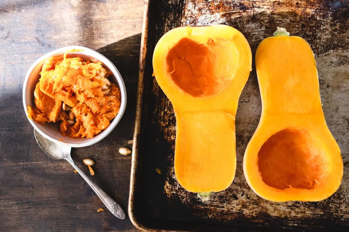Two butternut squash halves on a baking sheet next to bowl of seeds and pulp. 