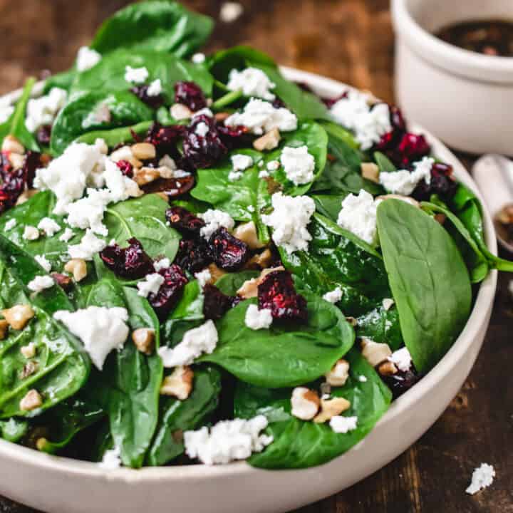Salad bowl of baby spinach, goat cheese crumbles, dried cranberries, and walnuts with bowl of dressing.