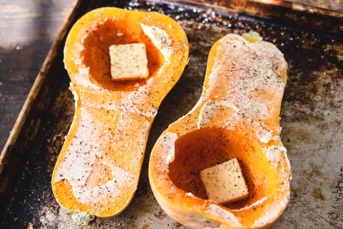 Two buttered and seasoned butternut squash halves on a baking sheet.