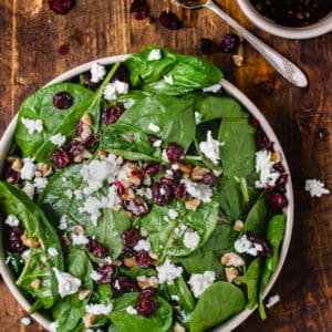 Spinach leaves, dried cranberries, walnut pieces and crumbled cheese in a bowl on wooden backdrop.