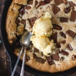 Cast iron skillet with a chocolate chunk cookie topped with vanilla ice cream scoops and two spoons.