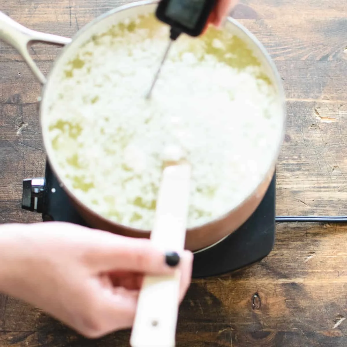 A pot of cheese curds with a thermometer and wooden spoon being heated.