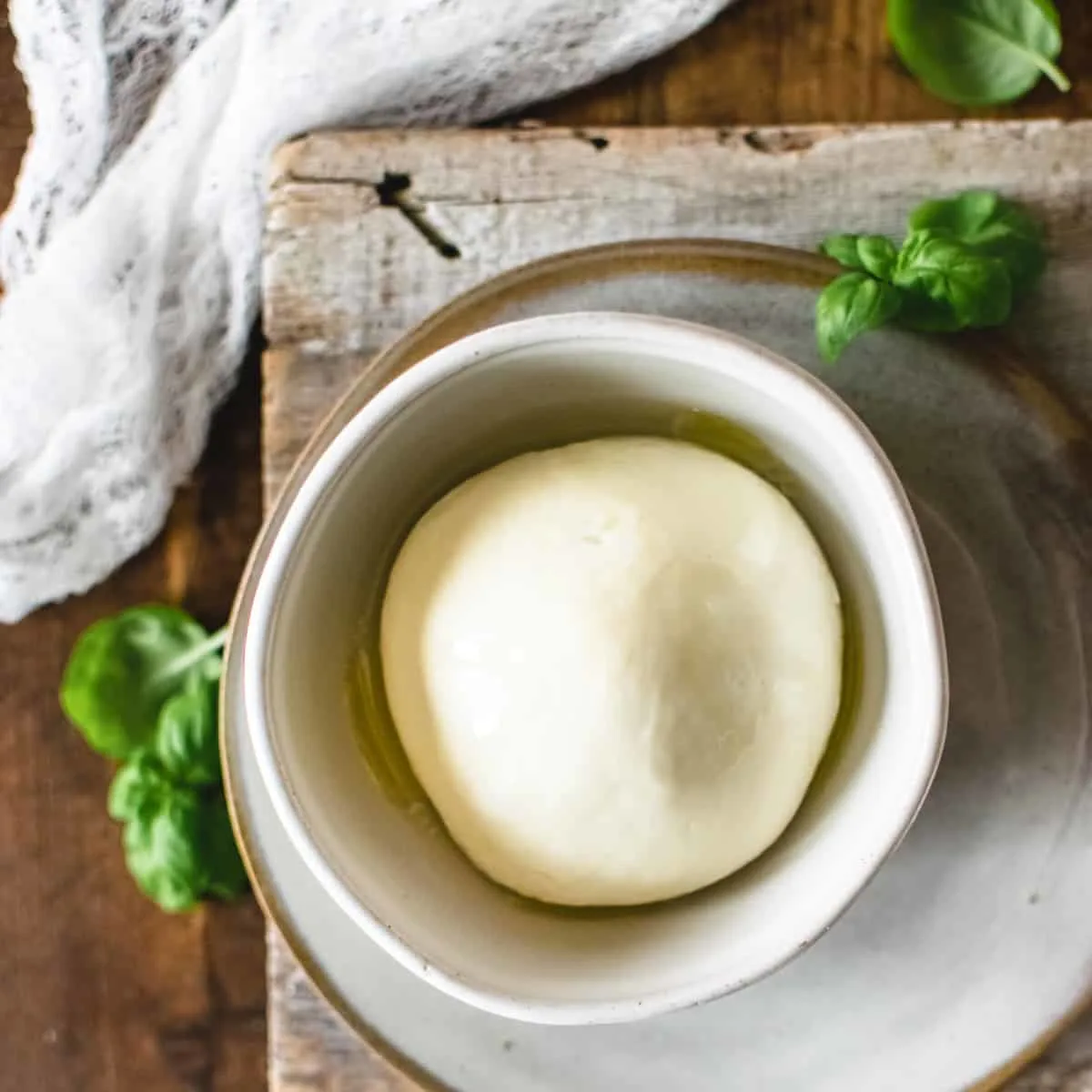 Ball of mozzarella cheese with basil and olive oil next to cheesecloth.