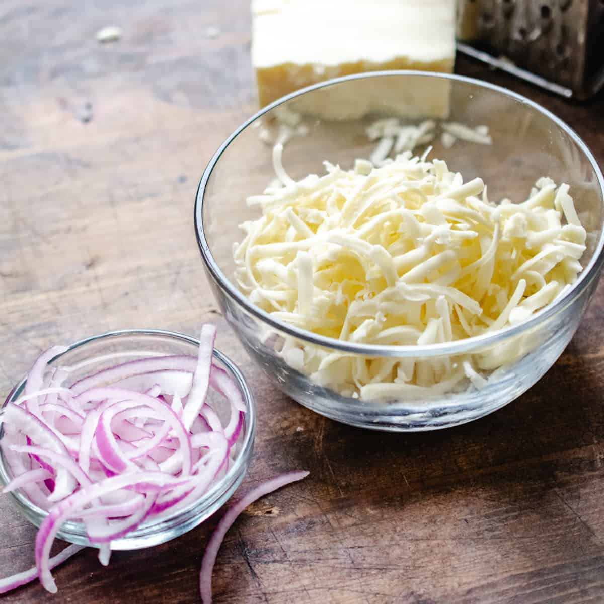 A glass bowl of sliced red onions and bowl of shredded mozzarella cheese and cheese grater on wooden surface.