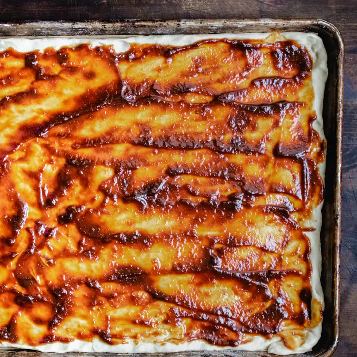 Flatbread dough covered with BBQ sauce on a baking sheet.