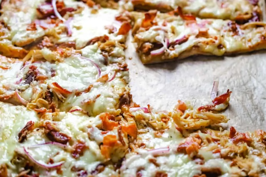 Close-up of a freshly baked pizza topped with melted cheese, shredded chicken, bacon bits, and thinly sliced red onions on a wooden cutting board. A slice has been removed, revealing the toppings and a crispy golden crust.