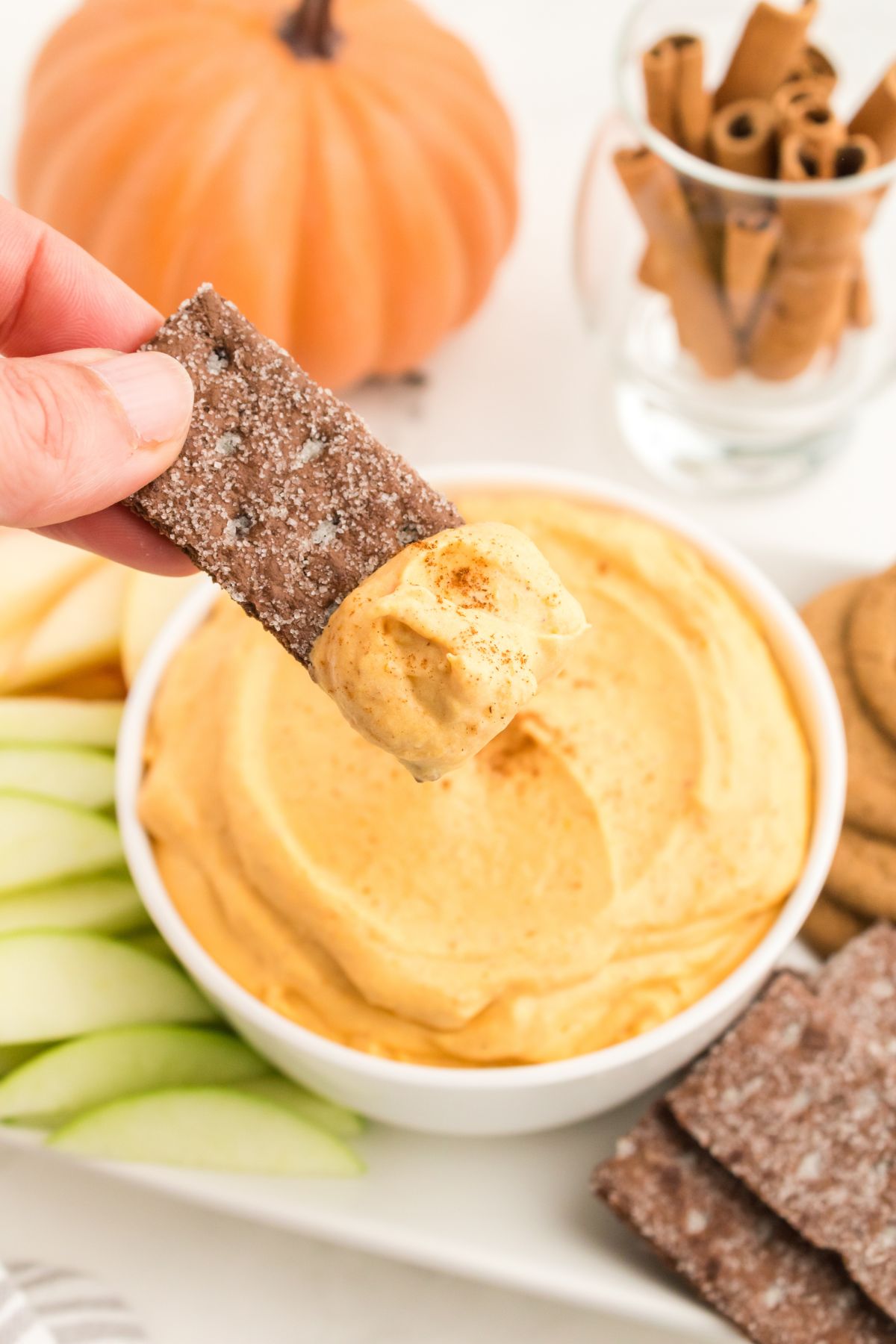A chocolate graham cracker dipped in Pumpkin Dip held above a bowl full of it.